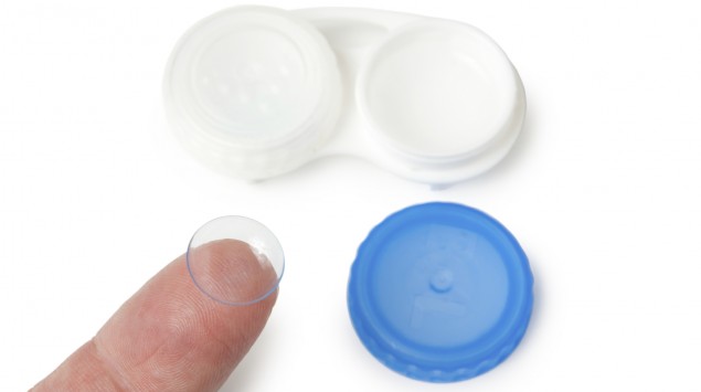 Auf einer Fingerspitze liegt eine Kontaktlinse, daneben steht eine geöffnete Kontaktlinsendose.
