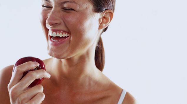Man sieht eine lachende junge Frau mit einem Apfel in der Hand.