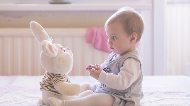 Das Bild zeigt ein Baby, das auf einer Decke sitzt und einen Kuschelhasen anschaut.