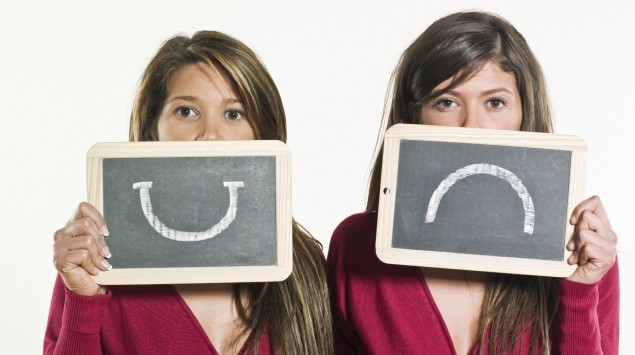 Zwei gleich gekleidete Frauen, eine hält ein Schild mit einem lachenden, die andere ein Schild mit einem traurigen Smiley hoch.