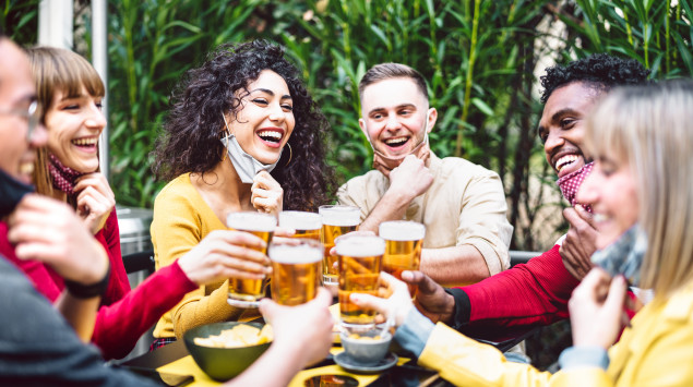 Junge Menschen stoßen im Biergarten miteinander an, die Masken runtergezogen