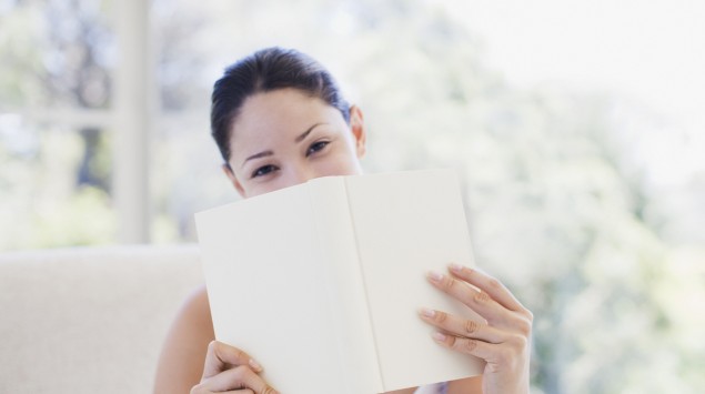 Eine junge lächelnde Frau hält ein Buch vors Gesicht, sodass ihr Mund verdeckt ist.