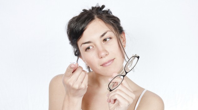 Eine Frau hält eine Brille in der linken Hand und betrachtet eine auf ihrem rechten Zeigefinger liegende Kontaktlinse.