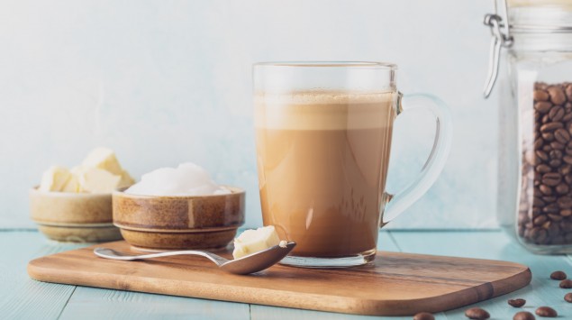 Auf einem Tisch stehen ein Henkelglas mit Buletproof-Coffee neben einem Glas Kaffee-Bohnen und einem Schälchen mit Butter.