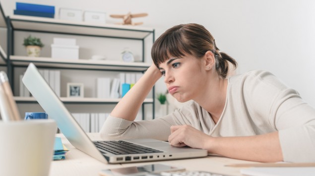 Eine junge Frau stützt den Kopf auf den Arm und starrt lustlos auf ihren Laptop.