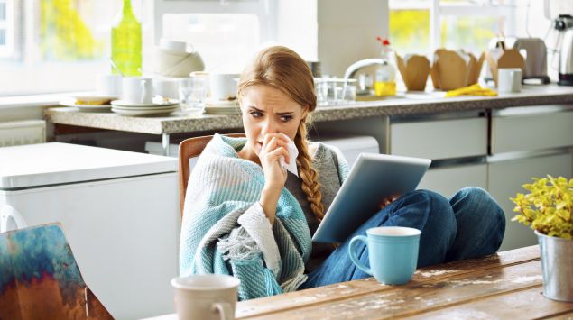 Kommen die Zahnschmerzen von der Erkältung? Eine verschnupfte Frau sitzt am Küchentisch mit einem Tablet und einer Tasse Tee.