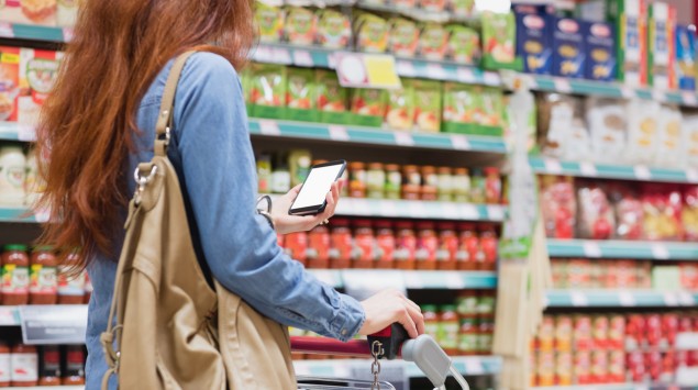 Eine Frau schiebt im Supermarkt einen Einkaufswagen vor sich her und schaut aufs Smartphone.