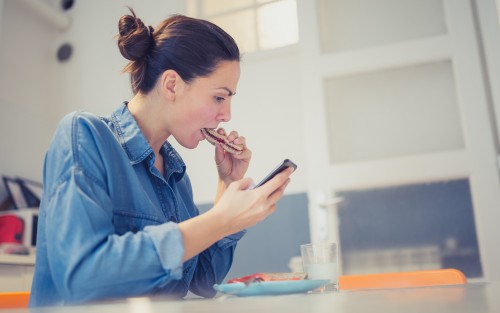 Eine Frau sitzt an einem Tisch und guckt auf ihr Smartphone, während sie ein Brot isst.