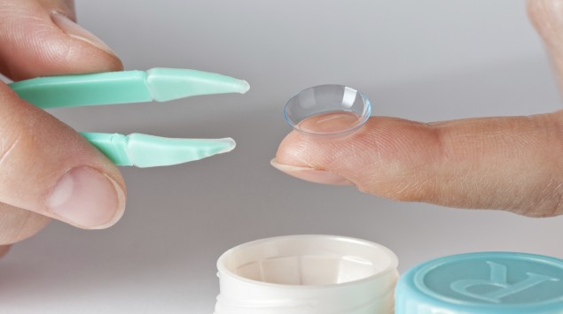 Die rechte Hand hält eine Kontaktlinsenpinzette, die linke eine weiche Kontaktlinse, darunter steht ein Kontaktlinsenbehälter.