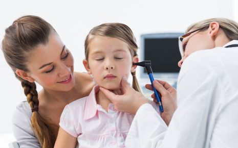 Eine Ärztin untersucht das Ohr eines Mädchen, das auf dem Schoß seiner Mutter sitzt.