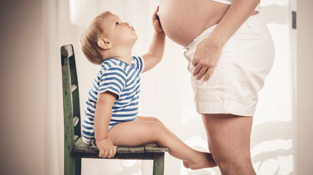 Das Bild zeigt ein Kleinkind, das auf einem Stuhl sitzt und den Bauch einer Schwangeren anfasst.
