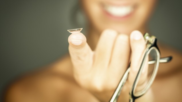 Eine lachende Frau hält eine Brille und eine Kontaktlinse in der Hand.