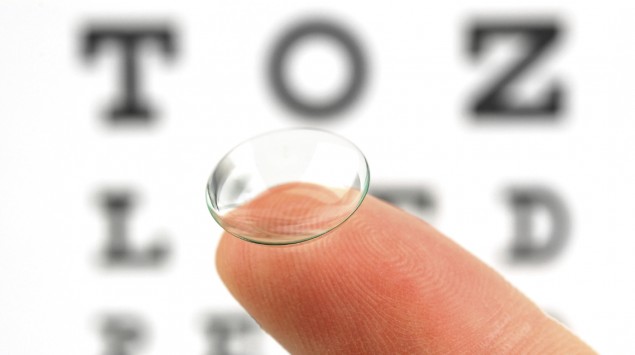 Eine Kontaktlinse liegt auf einer Fingerspitze, im Hintergrund ist eine Sehtesttafel zu sehen.