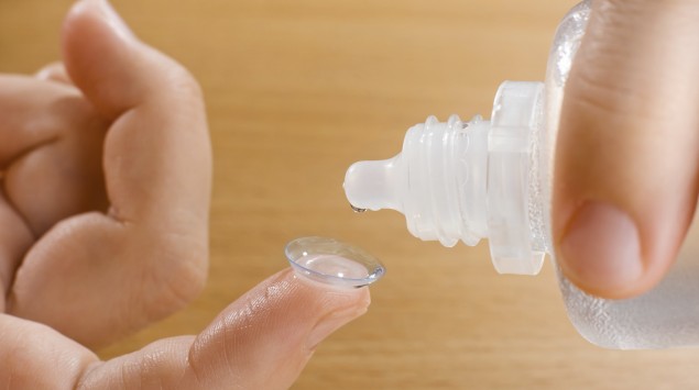 Zu sehen sind die Hände einer Person, die eine Lösung aus einer Plastikflasche auf eine Kontaktlinse träufelt.