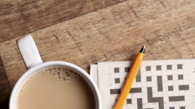 Das Bild zeigt einen Tischplatte, auf der ein Kaffee und ein Kreuzworträtsel liegt.