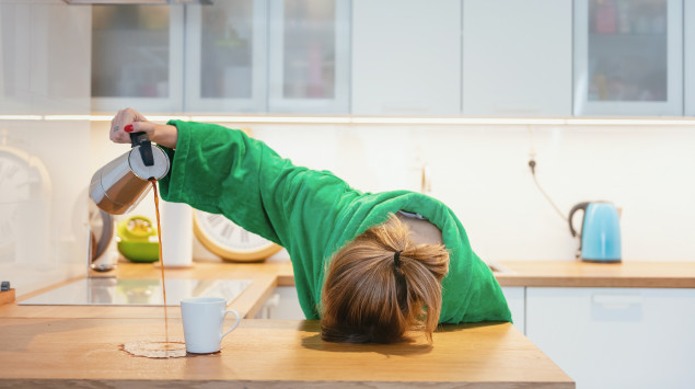 Eine Frau liegt mit dem Kopf auf dem Küchentisch, während sie den Kaffee neben die Tasse gießt