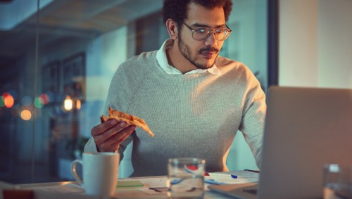 Ein Mann sitzt spätabends am Laptop und hält ein Stück Pizza in der Hand.