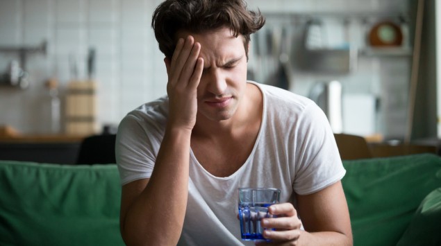 Man sieht einen Mann mit Kopfschmerzen und einem Glas Wasser in der Hand.