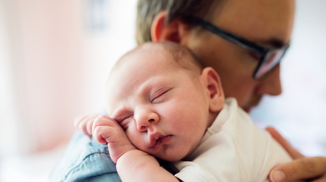 Ein junger Mann mit Brille trägt ein schlafendes Baby.
