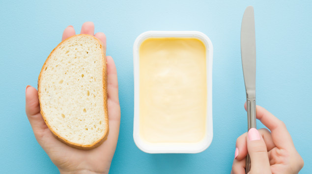 Man sieht zwei Hände, die eine Scheibe Brot und ein Messer halten, dazwischen ein Paket Margarine