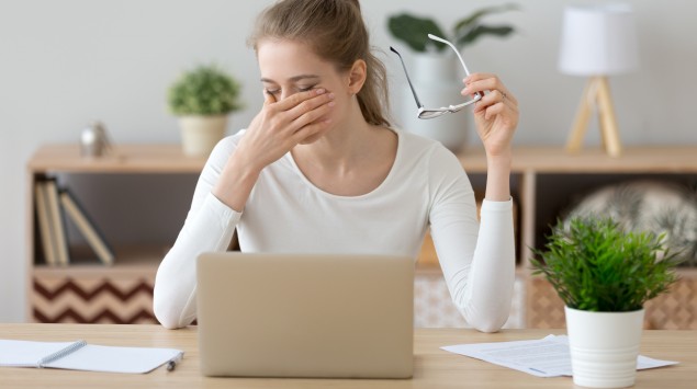 Eine Frau sitzt am Laptop, hat die Brille abgenommen und reibt sich die Augen.