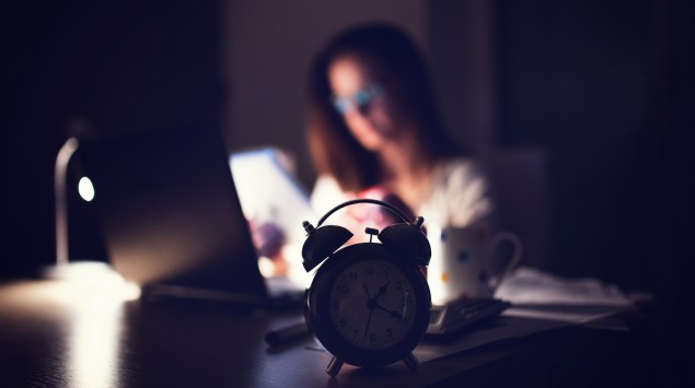 Auf einem Tisch steht ein Wecker, im Hintergrund ist eine junge Frau zu sehen, die im Schein einer Schreibtischlampe an einem Laptop arbeitet.