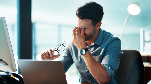 Ein junger Mann sitzt an einem Computerarbeitsplatz und reibt sich die Augen.