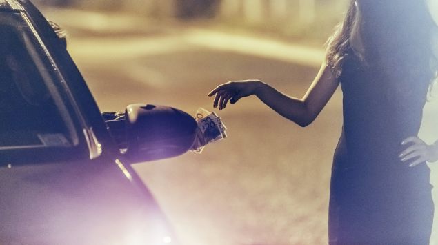 Im Dunkeln steht eine junge Frau neben einem Auto auf der Straße und nimmt vom Fahrer ein paar Geldscheine entgegen.