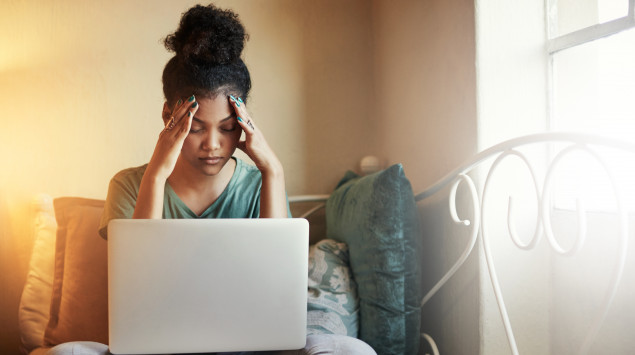 Eine Frau sitzt mit ihrem Laptop auf dem Bett und hält sich gestresst die Hände an den Kopf.