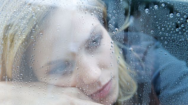 Eine depressiv aussehende Frau schaut durch ein mit Regentropfen bedecktes Fenster.