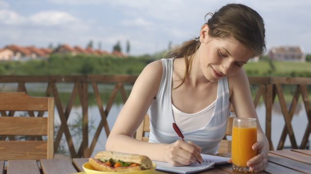 Das Bild zeigt eine Frau schreibend an einem Tisch mit ihrem Mittagessen vor ihr.