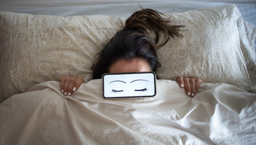 Eine Frau liegt im Bett und schläft, auf ihren Augen liegt ein Smartphone.