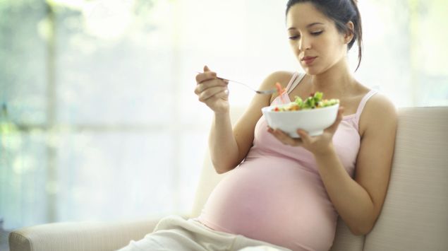 Eine schwangere Frau sitzt auf einem Sofa und isst Salat. Ausschließlich vegan ernähren sollten sich Schwangere jedoch besser nicht.