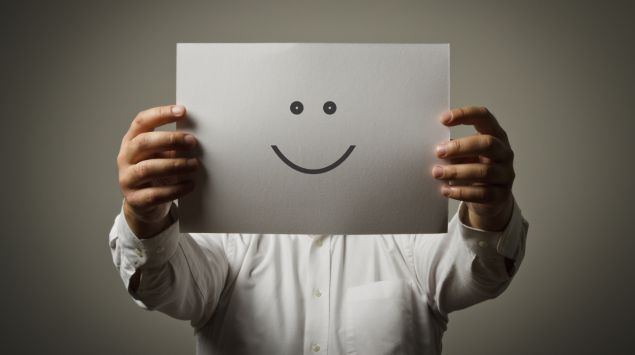 Ein Mann hält sich ein Blatt Papier mit einem aufgemalten Smiley vors Gesicht.