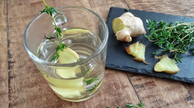 Auf einem Holztisch liegt ein Schneidebrett mit Thymianzweigen und angeschnittener Ingwerwurzel, daneben steht ein Glas mit Thymian-Ingwer-Tee.