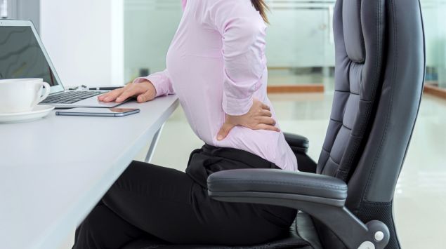 Eine in einem Bürostuhl sitzende Frau fasst sich an den verspannten Rücken.