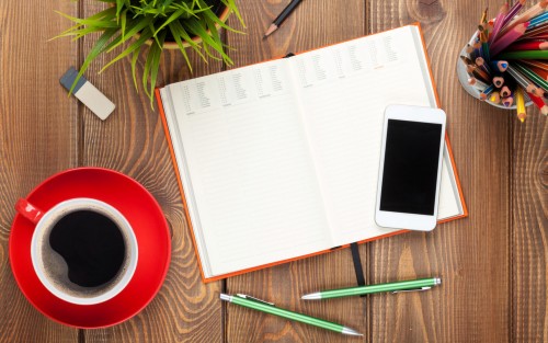 Ein offener Kalender, daneben ein Smartphone, Stifte und eine Tasse Kaffee